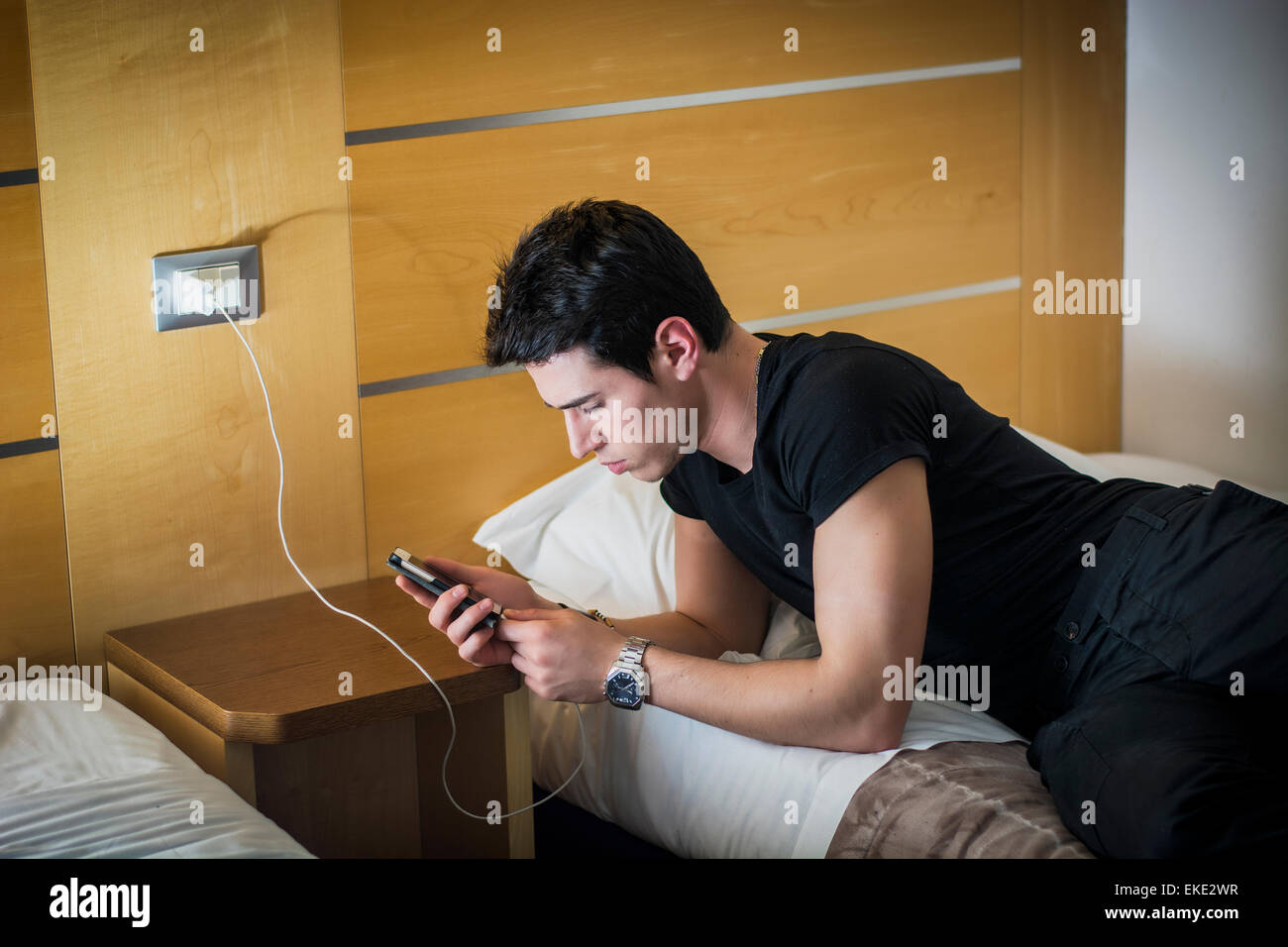 Serious Young Man Connecting a Phone to a Charger by His Bed Stock Photo