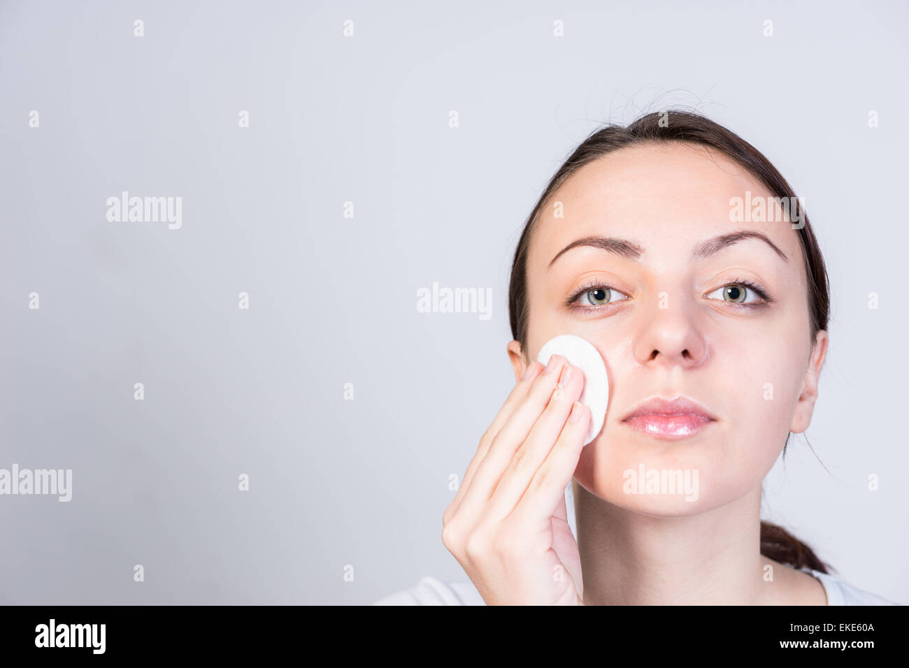 Close up Pretty Young Woman Scrubbing her Face with Astringent While Looking at the Camera. Captured in Studio on a Gray Background with Copy Space.. Stock Photo