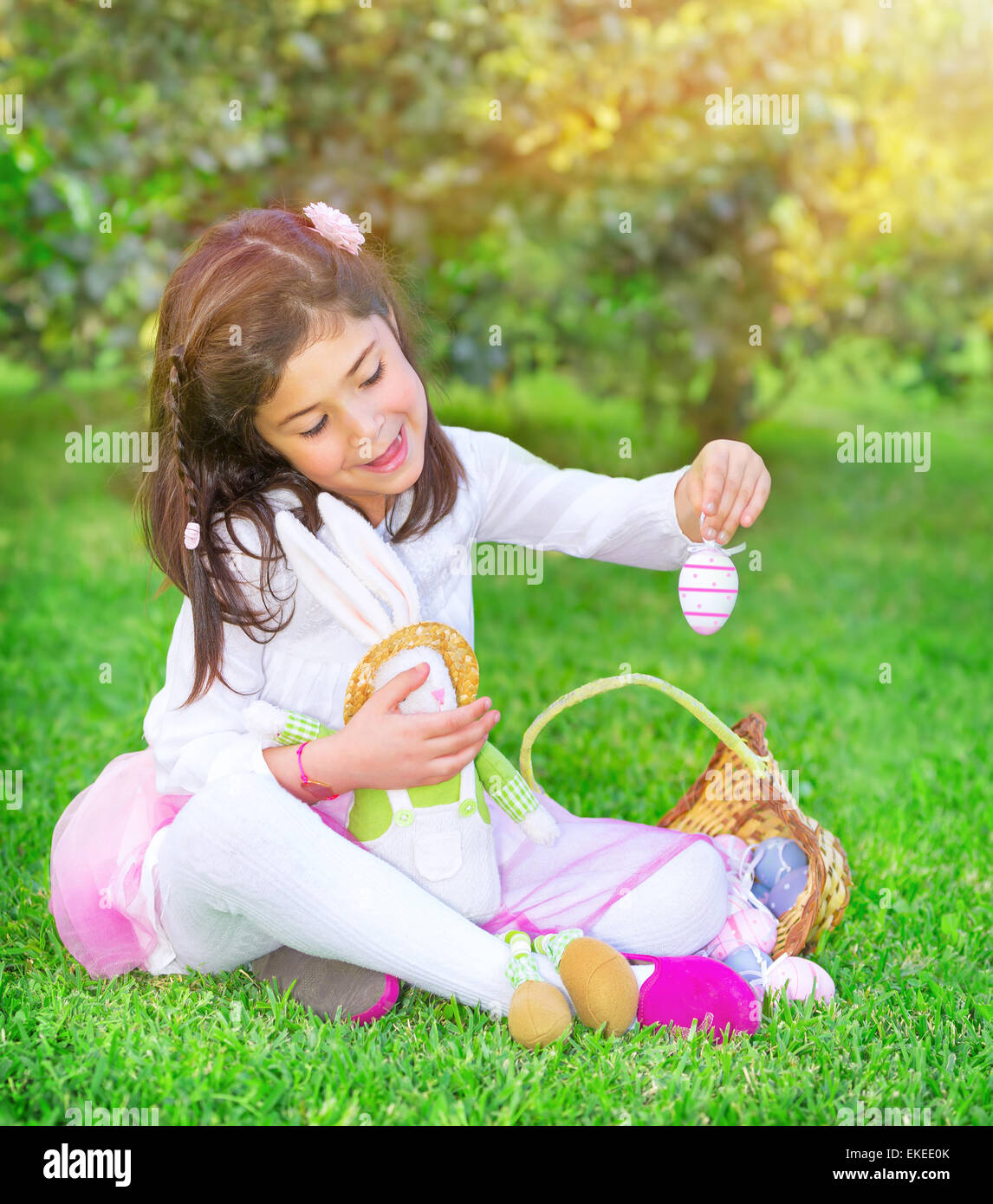 Cute little girl playing traditional Easter game, hunt for colored eggs, having fun in fresh green garden with cute little bunny Stock Photo