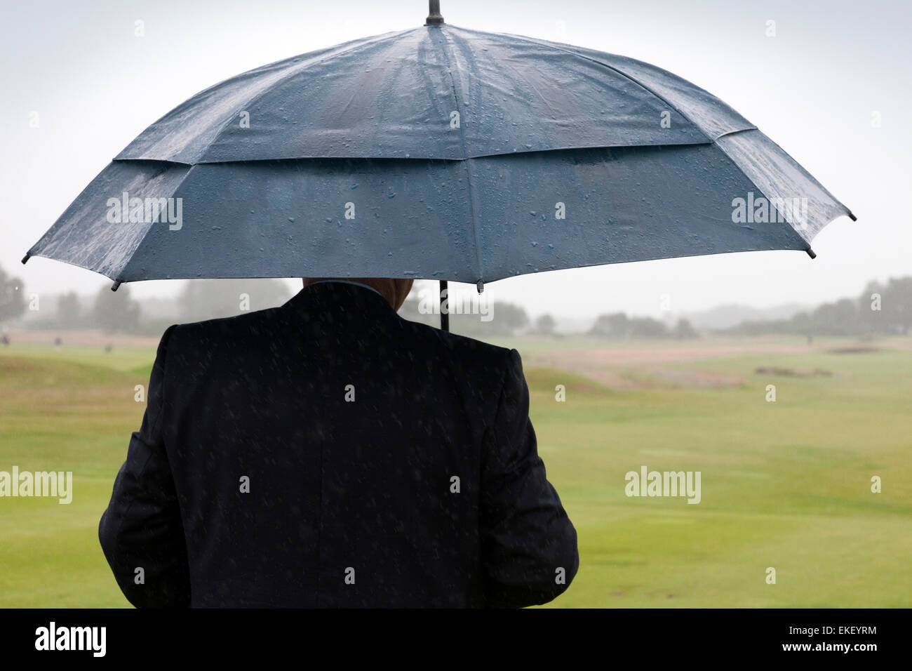 Golfer sheltering under an umbrella, looking out onto a golf course while it's raining Stock Photo