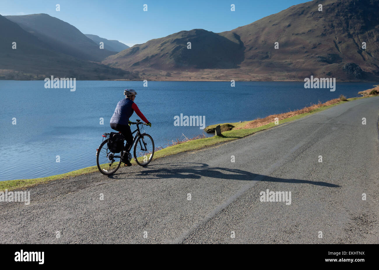 A female cyclist riding uphill on a small road alongside Crummock Water on a bright sunny day in Cumbria, England Stock Photo