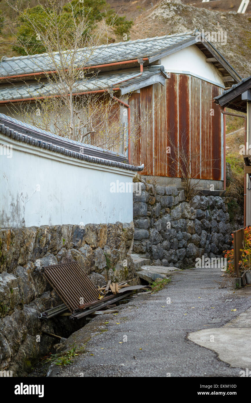 A rusted garage near a curving path and some trash against a white wall with traditional Japanese roof tiles. Stock Photo