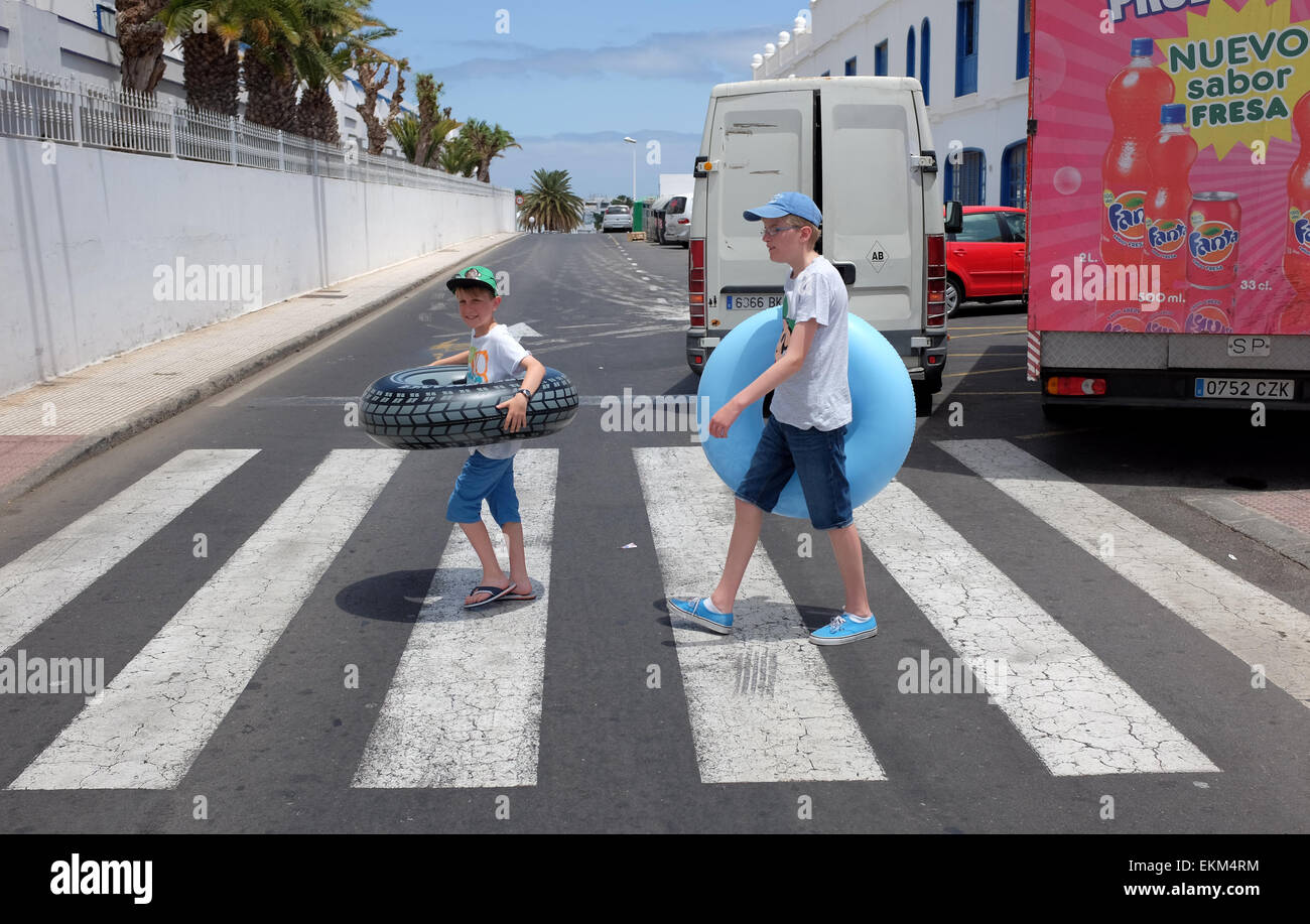 Two boys walk over a zebra crossing on their way to the beach with their rubber rings in Lanzarote Stock Photo