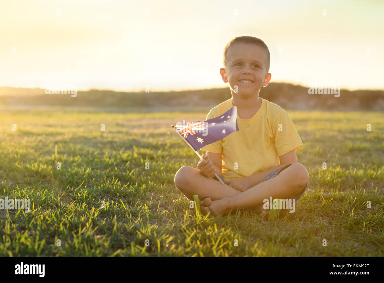 Cute smiling kid with flag of Australia sitting on the grass at sunset on Australia Day Stock Photo