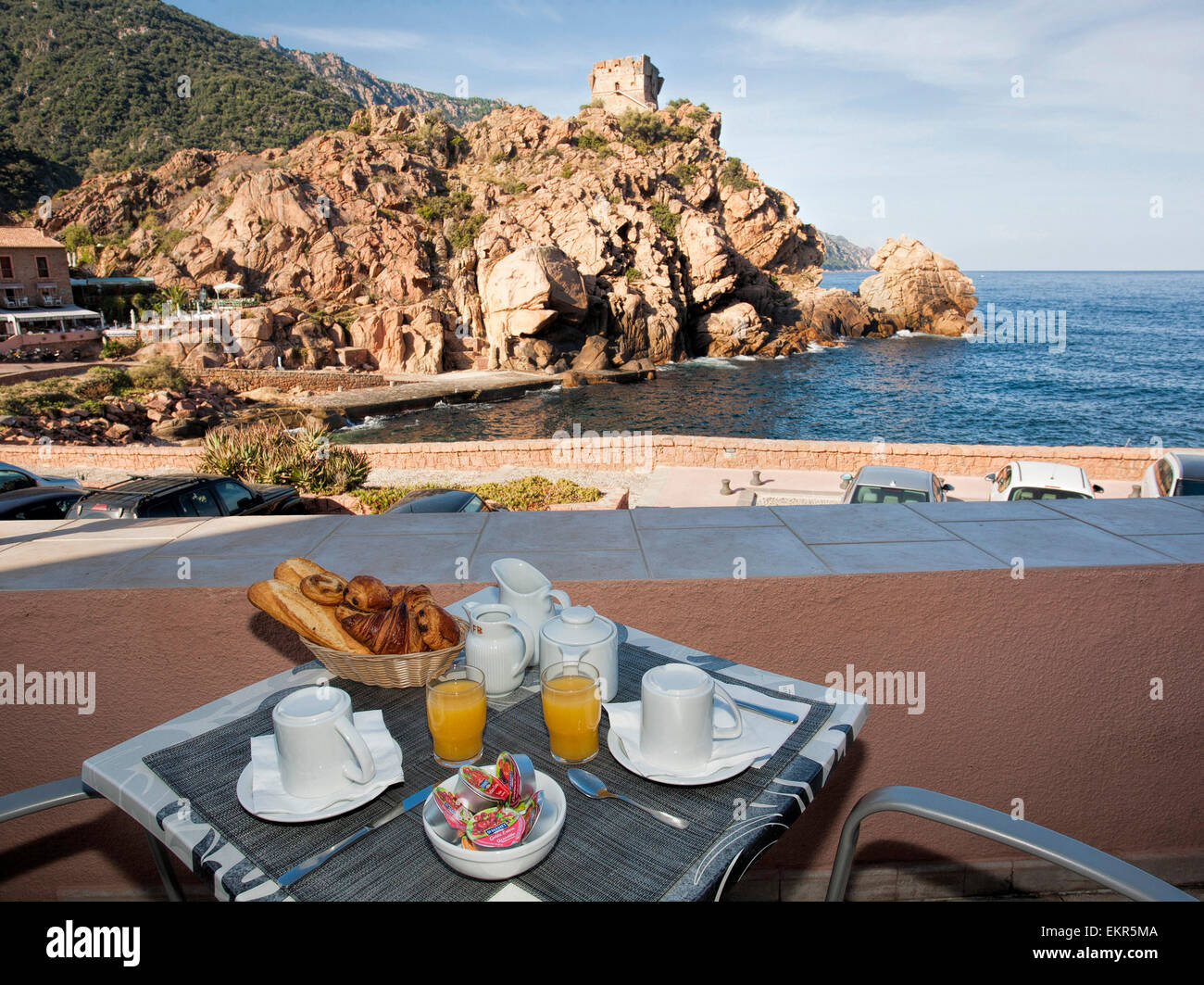 table with breakfast in Porto, Corsica, harbor, orange, bread, glass of orange juice, jam, milk, sea Stock Photo