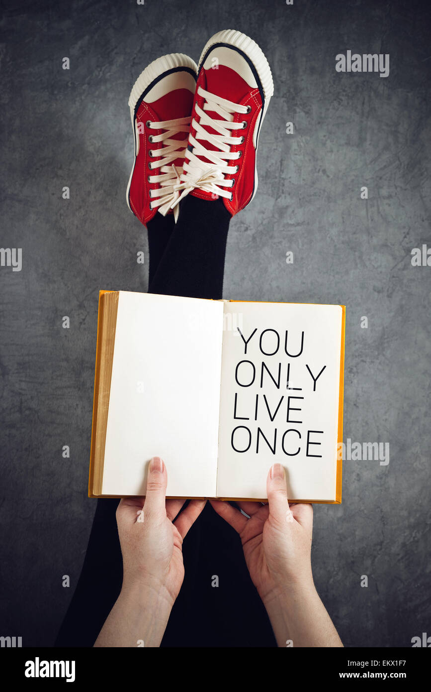 You Only Live Once or YOLO Concept with Young Woman Reading Book with Her Feet Raised in The Air Stock Photo