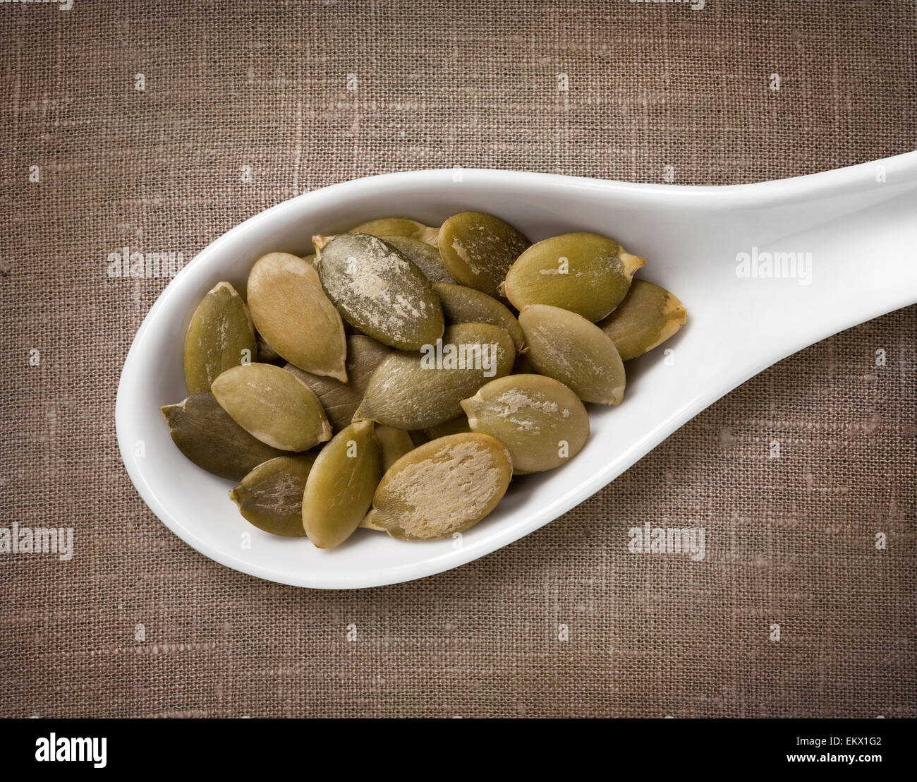 Peeled pumpkin seeds in white porcelain spoon / high-res photo of grain in white porcelain spoon on burlap sackcloth background Stock Photo