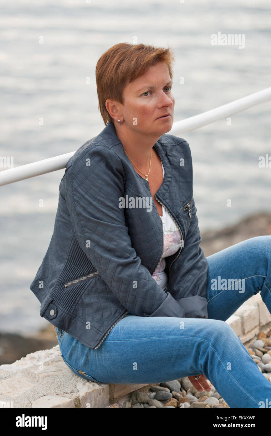 Middle aged caucasian tanned woman in autumn clothing sitting on the rocky sea beach Stock Photo
