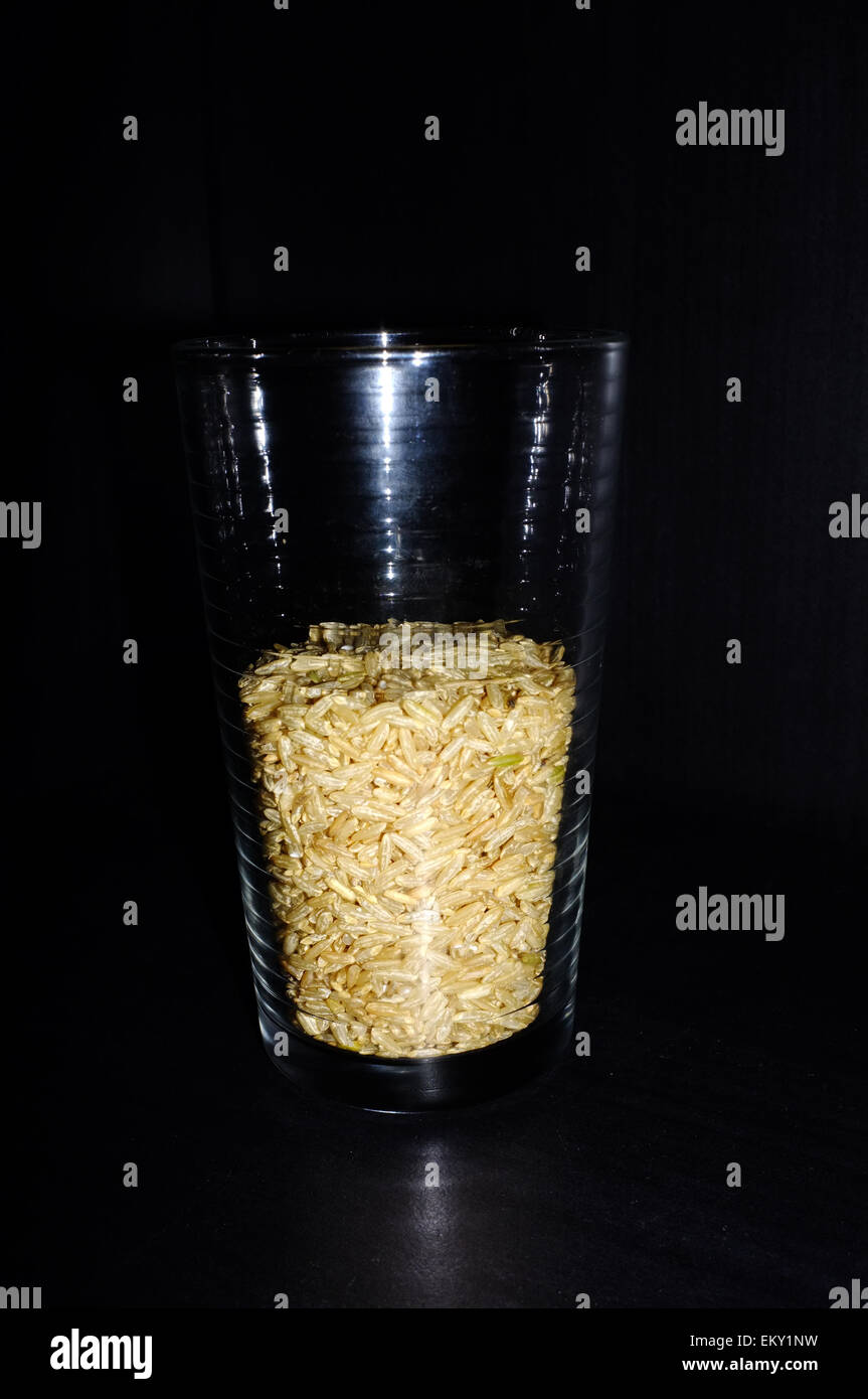 A glass half full/half empty of brown grain rice against a black background. Stock Photo