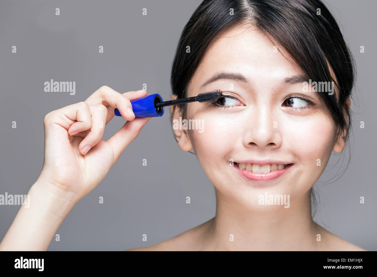 Chinese woman putting mascara on eyelashes on gray background Stock Photo