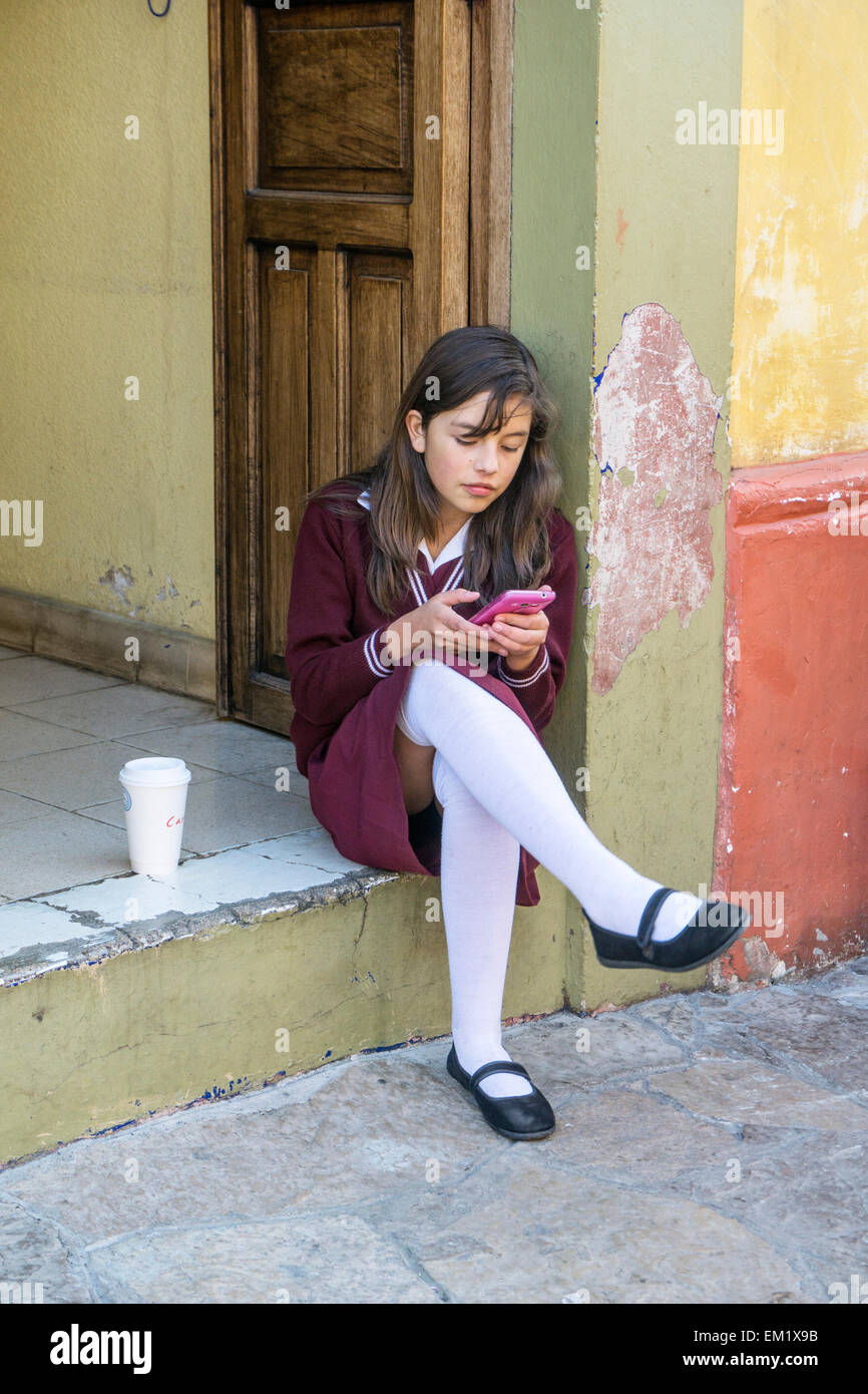 pretty teenage schoolgirl sits on stoop in school uniform with spotless white stockings using her cellphone San Cristobal Stock Photo