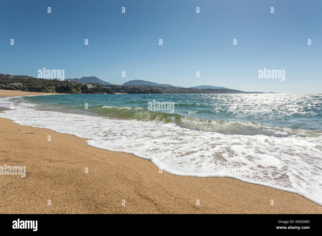 Waves gently lapping onto the beach at Propriano with golden sand, blue sea and blue sky and hills in the background Stock Photo