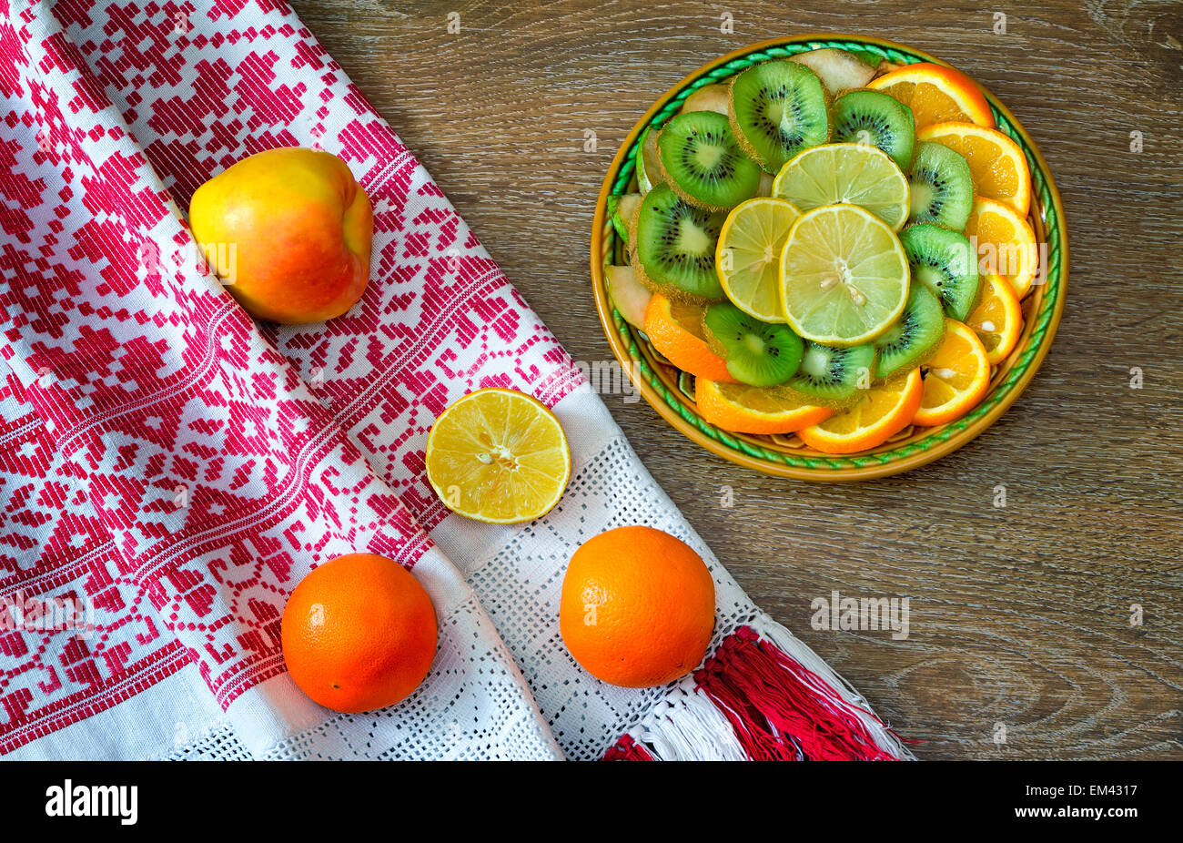 On the table is a dish of sliced fruits: oranges, kiwi, lemon. Near them on a beautiful towel are two oranges and an Apple. Stock Photo