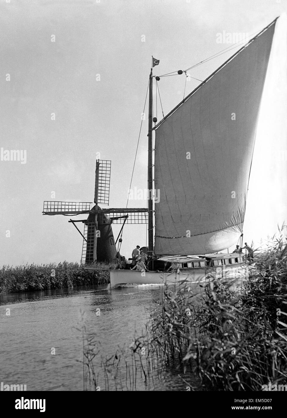 A traditional Norfolk Broad yachts sailing past one of the many windmill water pumps that help with the drainage of the Broads Landscape Sailing Circa 1935. Stock Photo