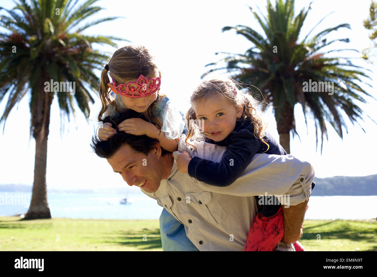 Mature man giving piggy back to daughter and friend in coastal park, New Zealand Stock Photo