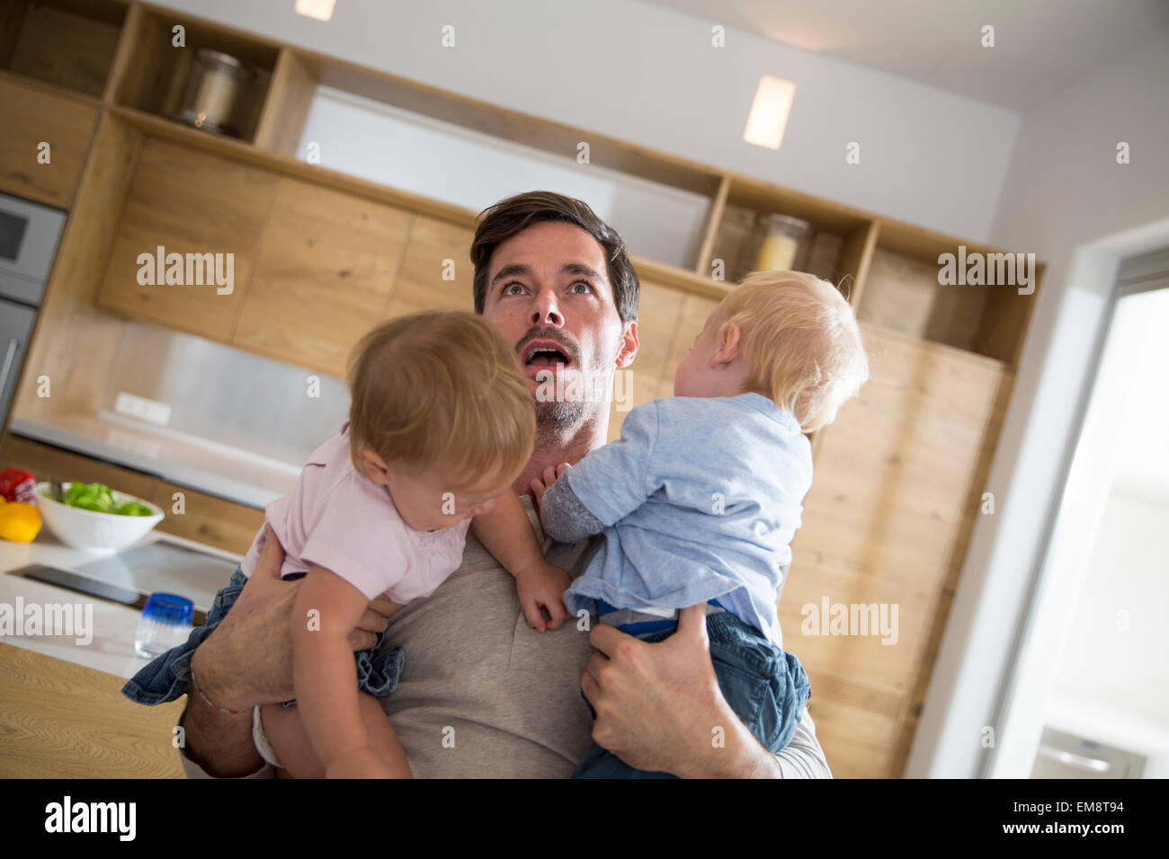 Father struggling to carry male and female twin toddlers in dining room Stock Photo