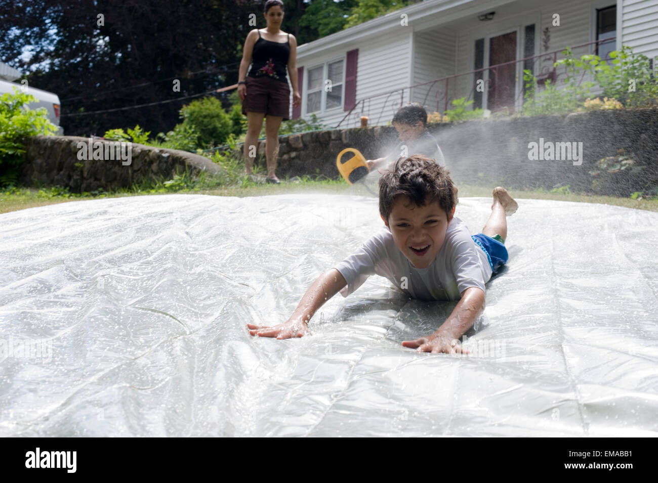 Seven year old boy plays on a homemade slip and slide in his backyard with his mother and 4 year old brother Stock Photo