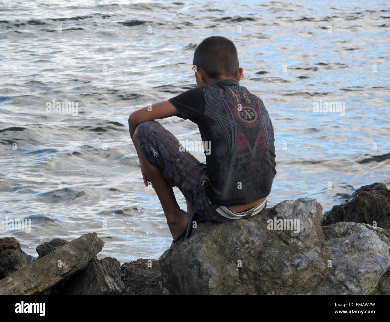 Back view of boy sitting on rock looking into water. Stock Photo