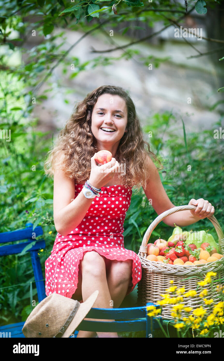 Smiling young woman with with wire basket of different fruits in a ...