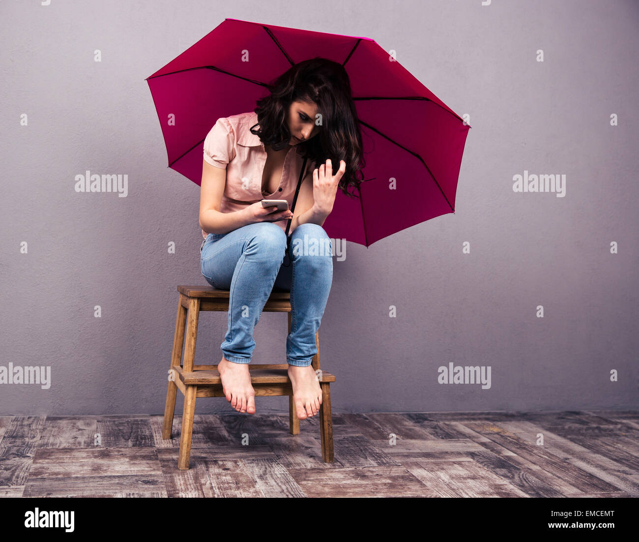 Young woman sitting on the chair with smartphone and pink umbrella. Wearing in shirt and jeans. Stock Photo