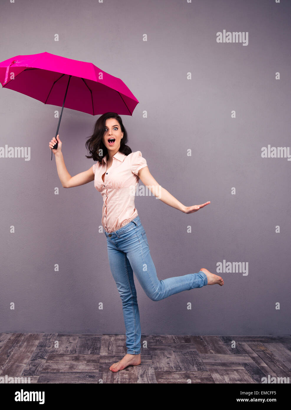 Full length portrait of a surprised woman posing with pink umbrella in studio. Wearing in jeans and shirt. Barefoot. Looking at  Stock Photo