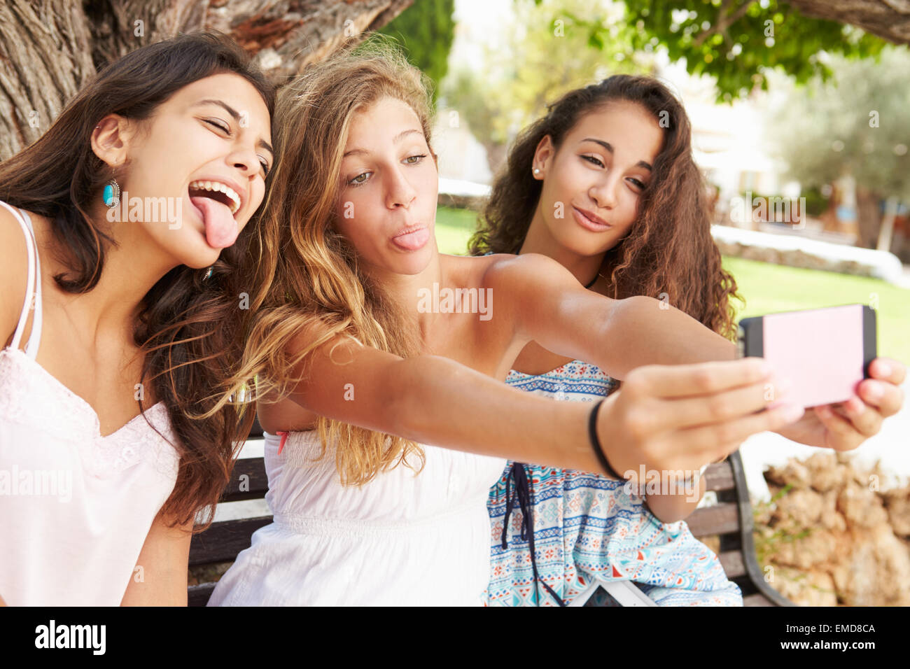 Three Teenage Girls Sitting On Bench Taking Selfie In Park Stock Photo