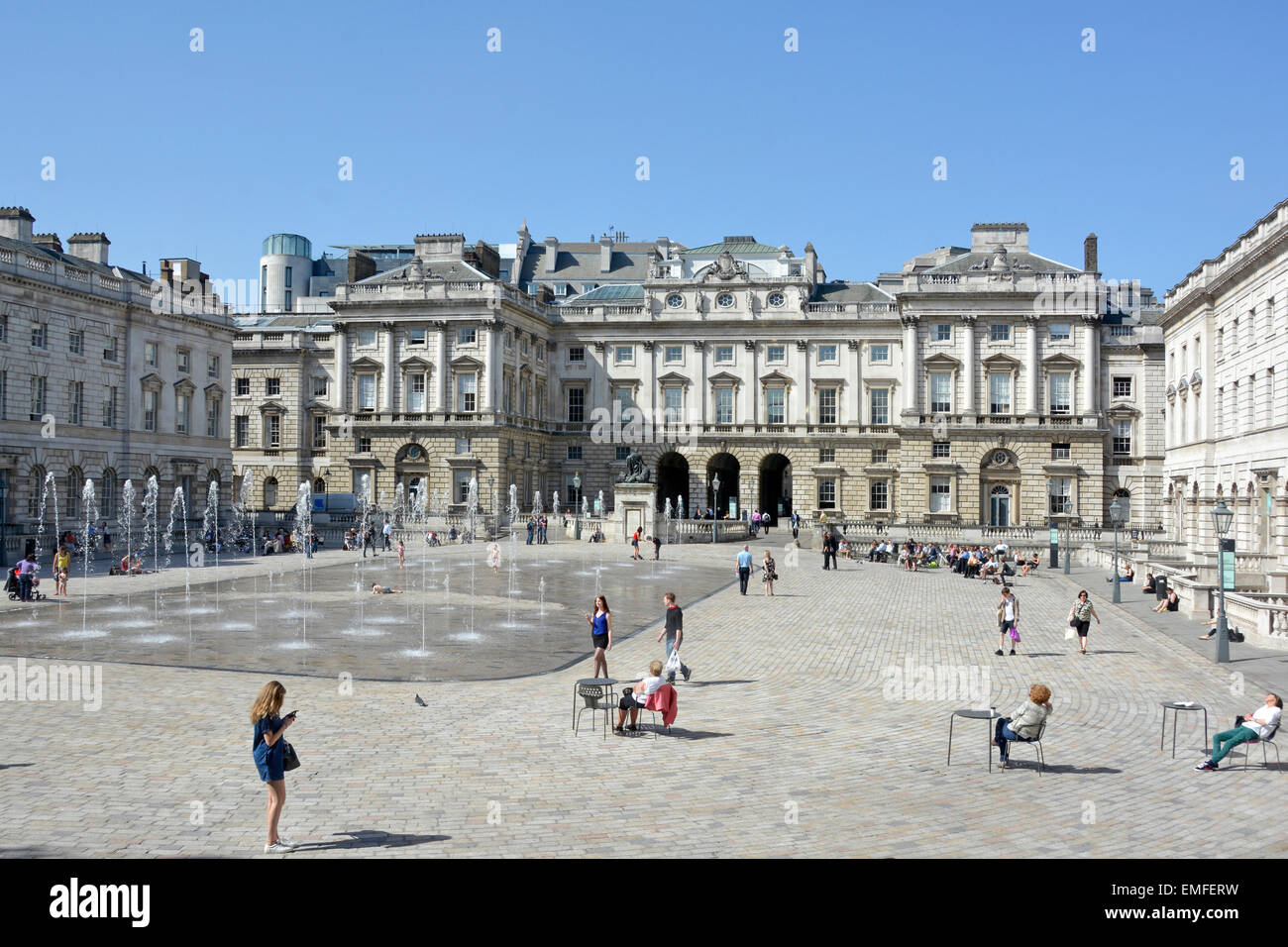People tourists enjoy fountains & Neoclassical buildings surrounding  cobblestone courtyard spring sunshine at Somerset House Strand London England UK Stock Photo