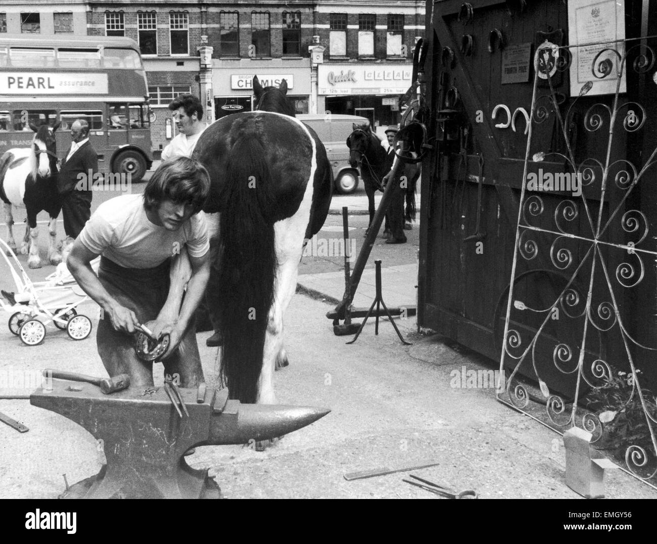 23 year old Londoner Derek Dewdney the village blacksmith of Brent, London, shoes the horses at his forge against a back cloth of a teeming street as tankers, trucks and buses thunder by. 30th July 1970 Stock Photo