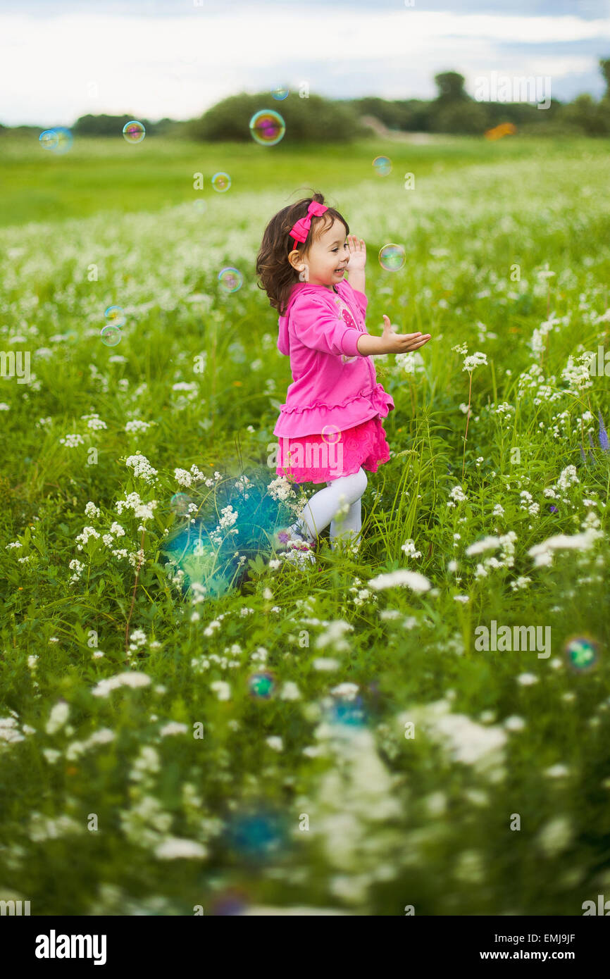 beautiful carefree girl playing outdoors in field with high green grass. little child running away from bubbles and laughing Stock Photo
