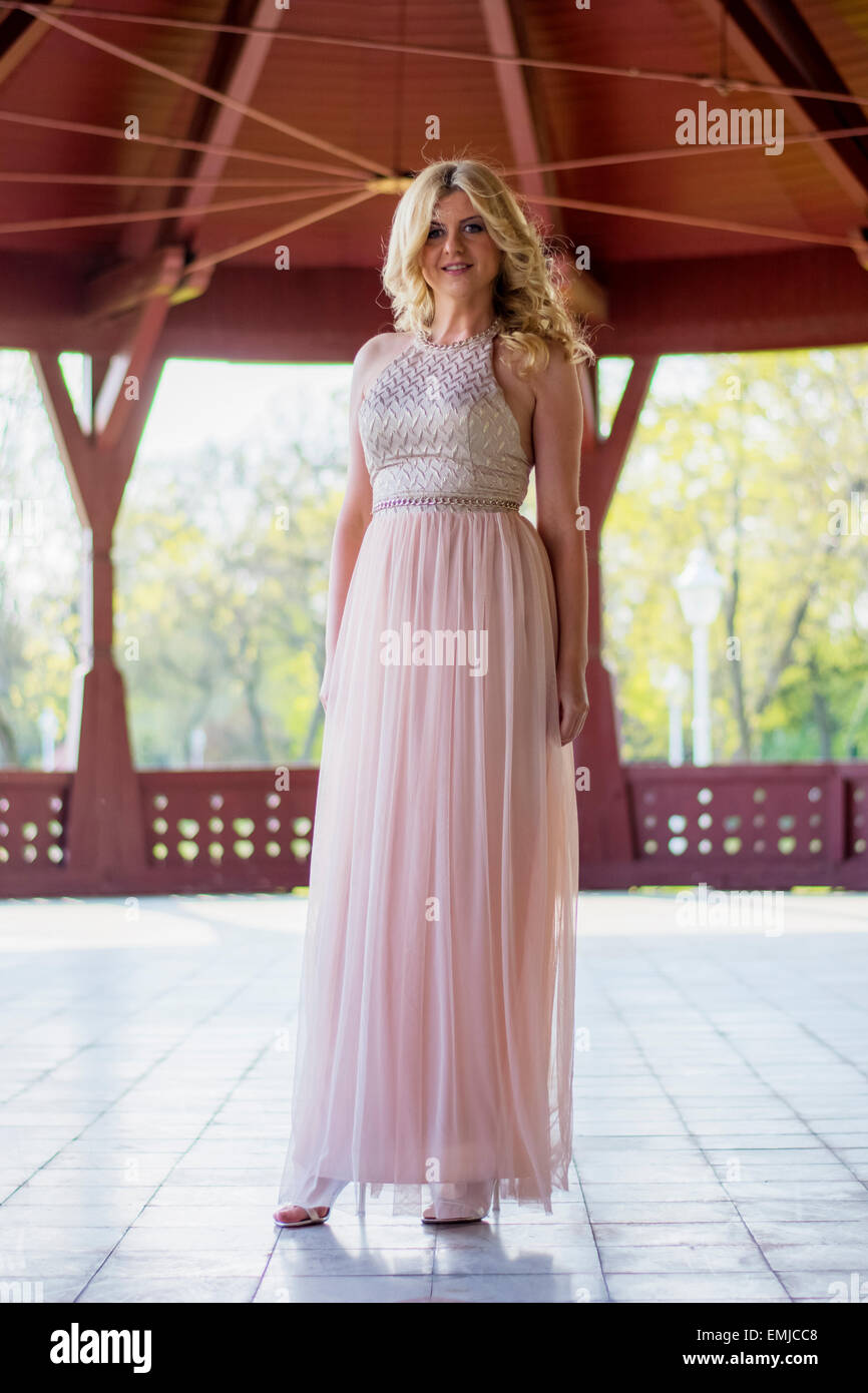Beautiful blond curly woman wearing evening peach color gown posing outdoors. Fashionable and glamorous dress and style. Stock Photo