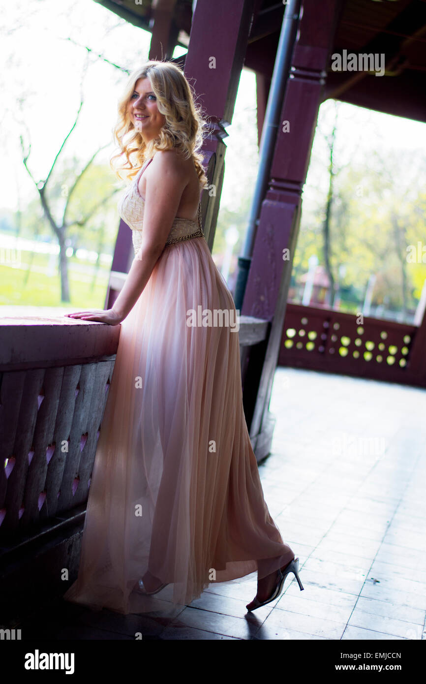 Beautiful blond curly woman wearing evening peach color gown posing outdoors. Fashionable and glamorous dress and style. Stock Photo