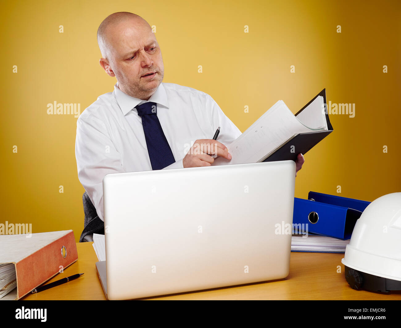 Hardworking male engineer in office, he wearing a white shirt and tie, the laptop, binders and white hard hat is on the table Stock Photo