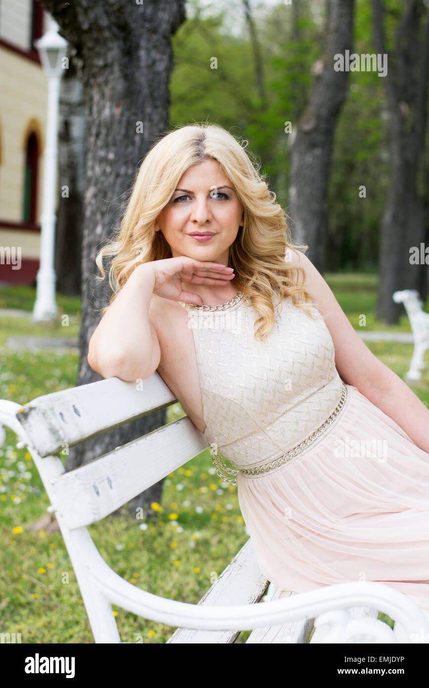Beautiful blond curly woman wearing evening peach color gown sitting on white bench outdoors. Fashionable and glamorous dress Stock Photo