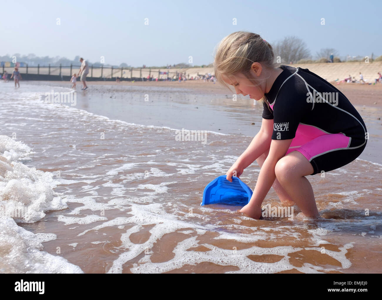 A young girl in a wetsuit collecting water with a bucket on the beach UK Stock Photo