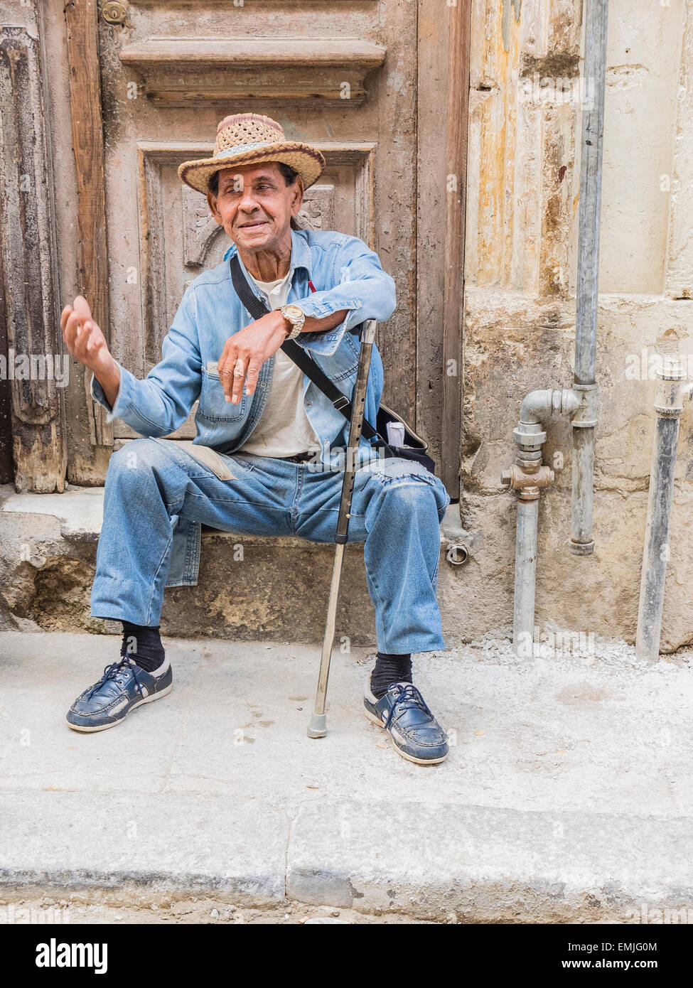 A Cuban male senior citizen gestures with his right hand as he leans on his cane and sits on a concrete step by his house. Stock Photo