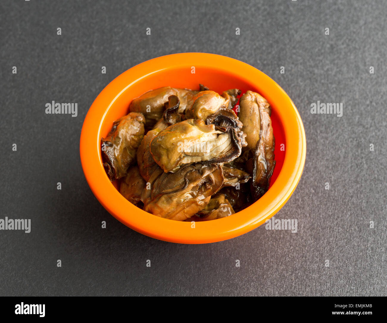 Several small whole smoked oysters in a bright orange bowl atop a black table top. Stock Photo