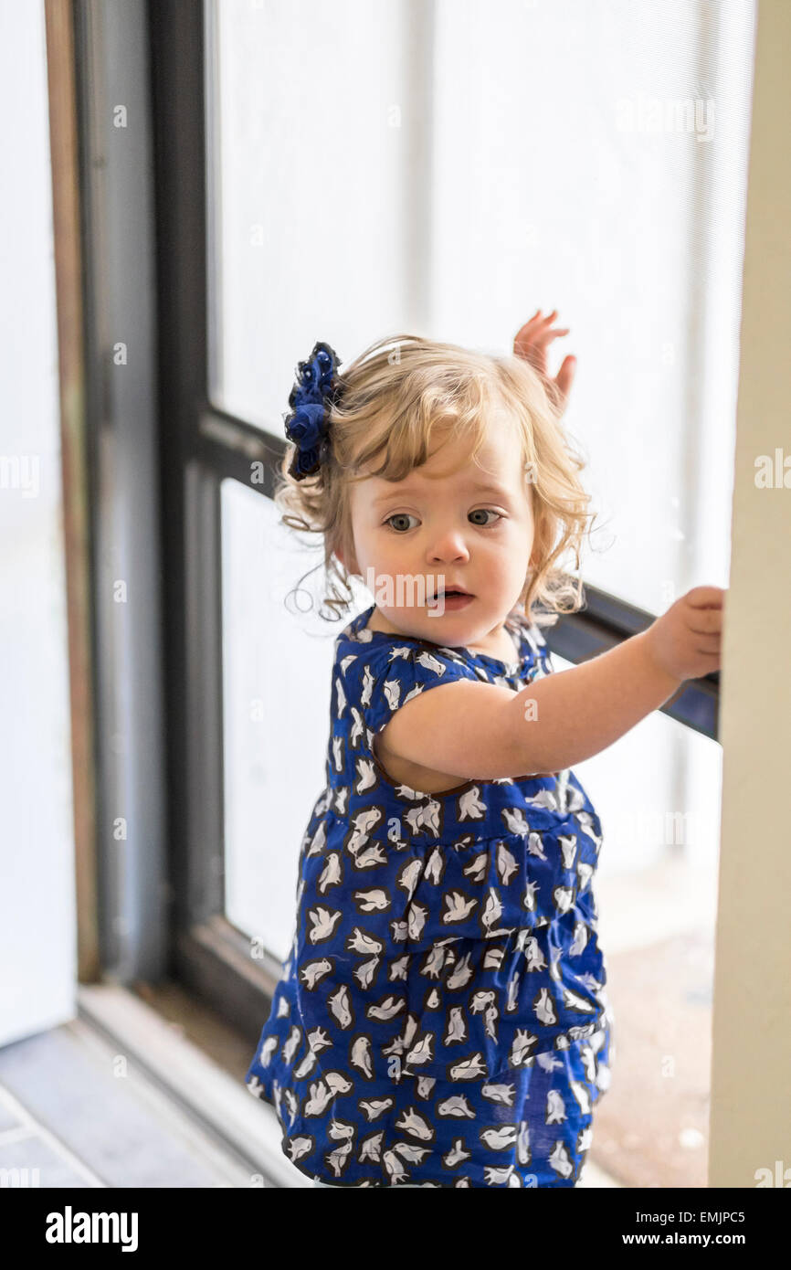 A beautiful 14 month old Caucasian girl stands near a door with lovely north light looking at camera. Stock Photo