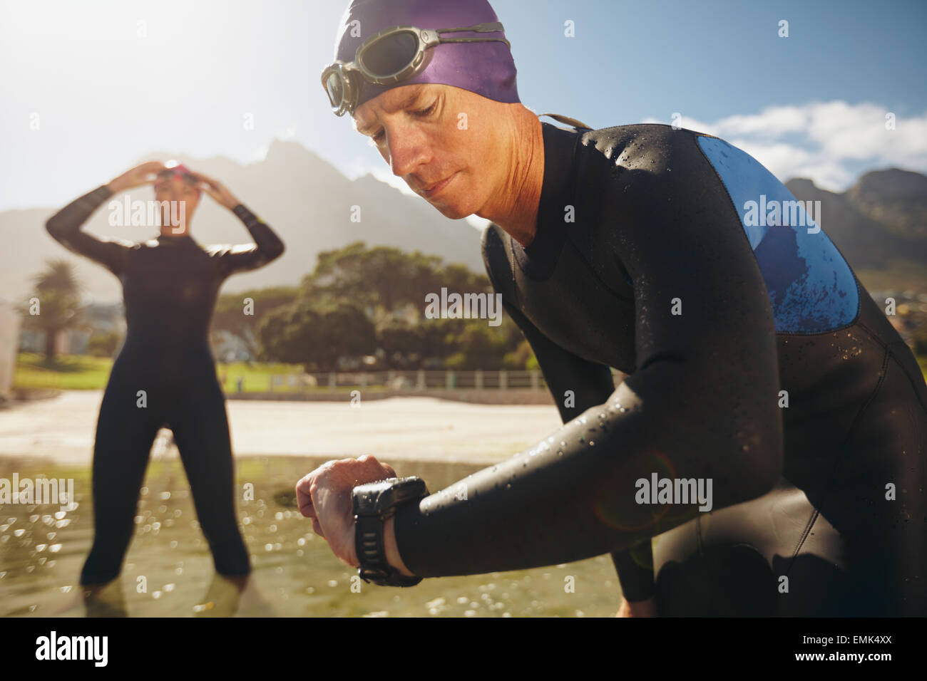 Focused young man checking his watch while in wetsuit at the lake. Man looking at watch after practice run. Triathletes. Stock Photo