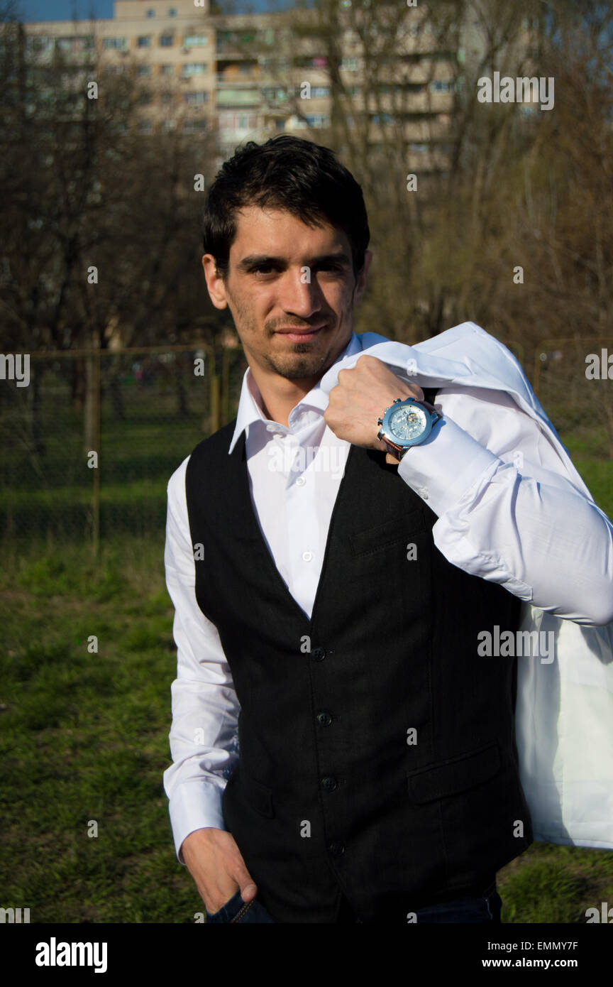 Young handsome and fashion man with straight hair outside  wearing a black vest, white shirt, wristwatch, looking at the camera Stock Photo