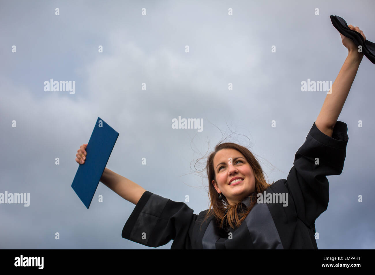 Pretty, young woman celebrating joyfully her graduation - spreading wide her arms, holding her diploma, savouring her success (c Stock Photo