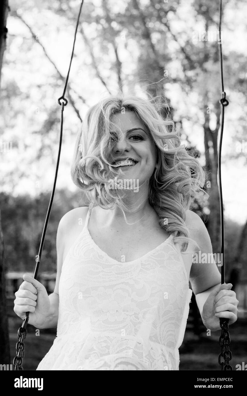 Smiling blond curly woman wearing white lace dress enjoying swing. Monochrome effect. Stock Photo