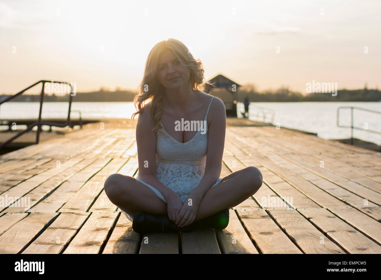 Portrait of a blond woman sitting on a deck wearing white lace dress and short black boots with lake behind her enjoying sun. Stock Photo