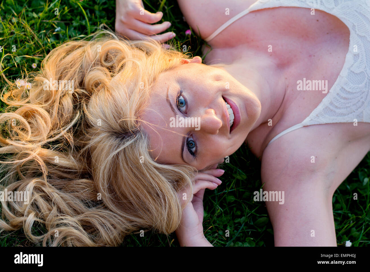 Portrait of a blond curly woman laying on a grass wearing white lace dress enjoying sunset. Stock Photo