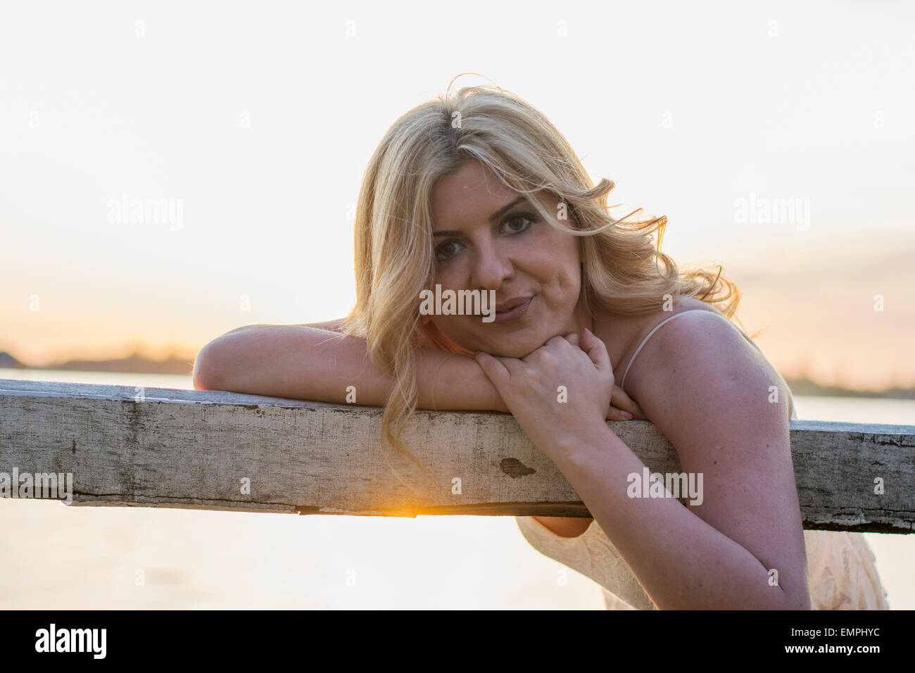 Portrait of beautiful blond curly woman sitting on old wooden bench with lake behind. Stock Photo