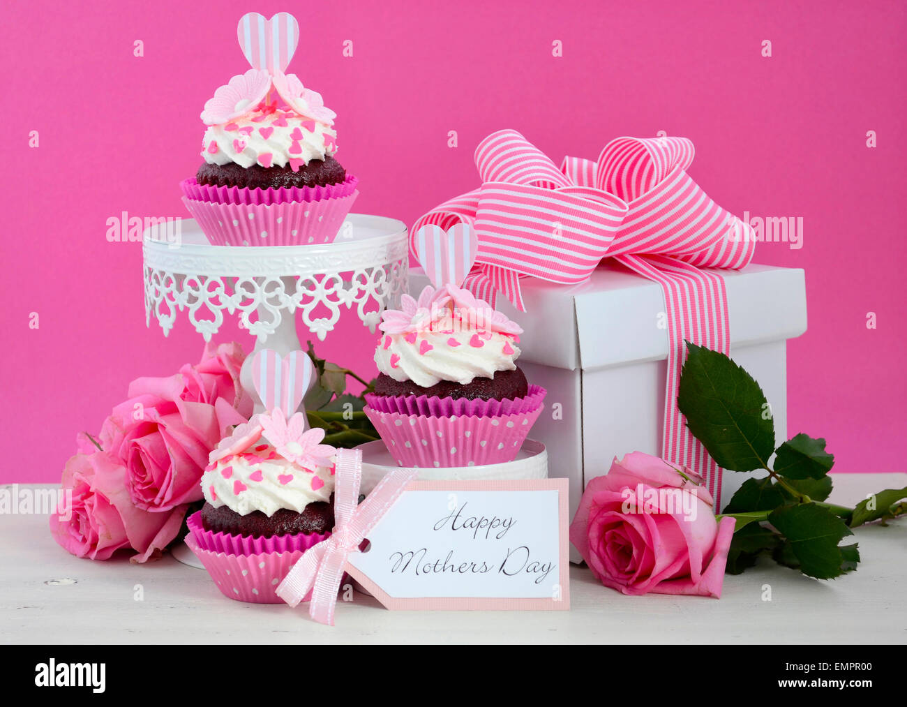 Happy Mothers Day pink and white cupcakes on retro style cake stands and large gift box on vintage white wood table. Stock Photo