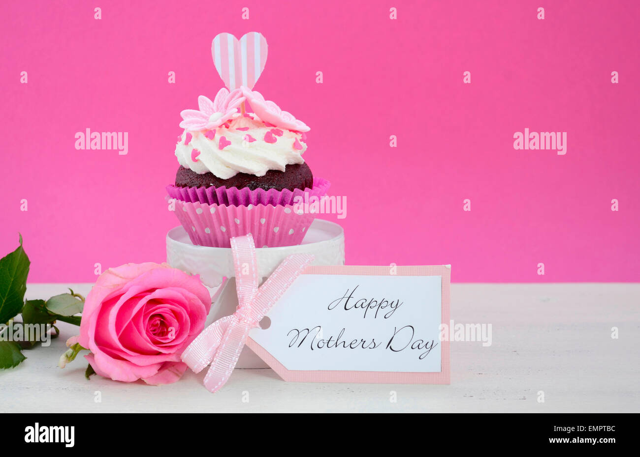 Happy Mothers Day pink and white cupcake on retro style cake stand and pink rose on vintage white wood table, with copy space. Stock Photo