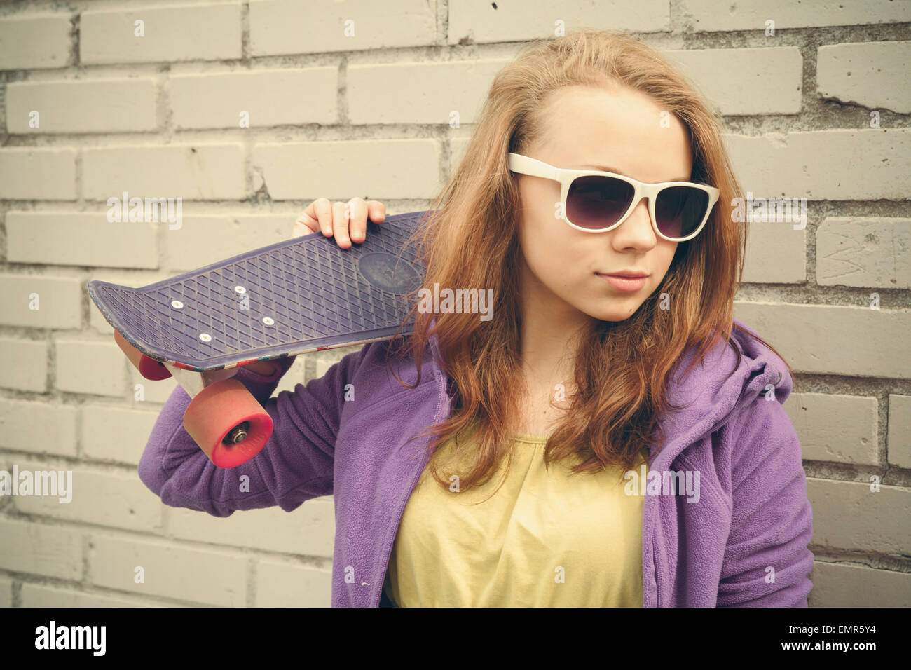 Blond teenage girl in sunglasses holds skateboard near gray urban brick wall, vintage soft tonal correction, old style photo Stock Photo