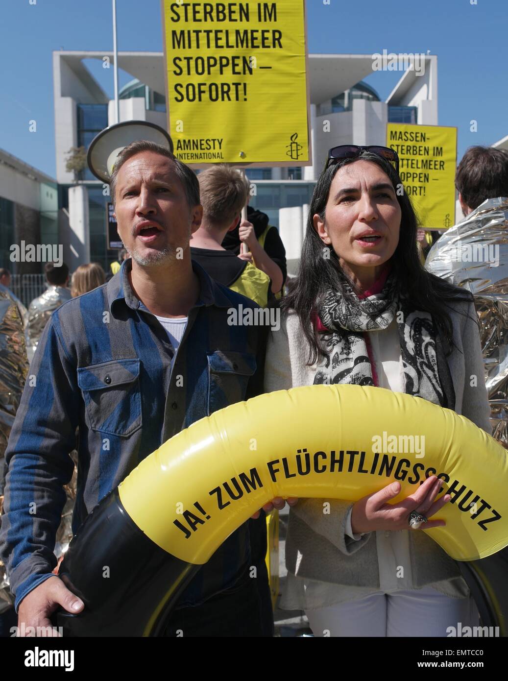 Berlin, Germany. 23rd Apr, 2015. Actor Benno Fuermann and Amnesty secretary general, Selmin Çaliskan, demonstrate under the slogan 'Save the refugees - Now!' with a life preserver written with 'Yes! to refugee protection!' in front of the Federal Chancellery in Berlin, Germany, 23 April 2015. In the background a demonstrator holds up a sign that reads 'Stop mass deaths in the Mediterranean - At once!' Photo: JOERG CARSTENSEN/dpa/Alamy Live News Stock Photo