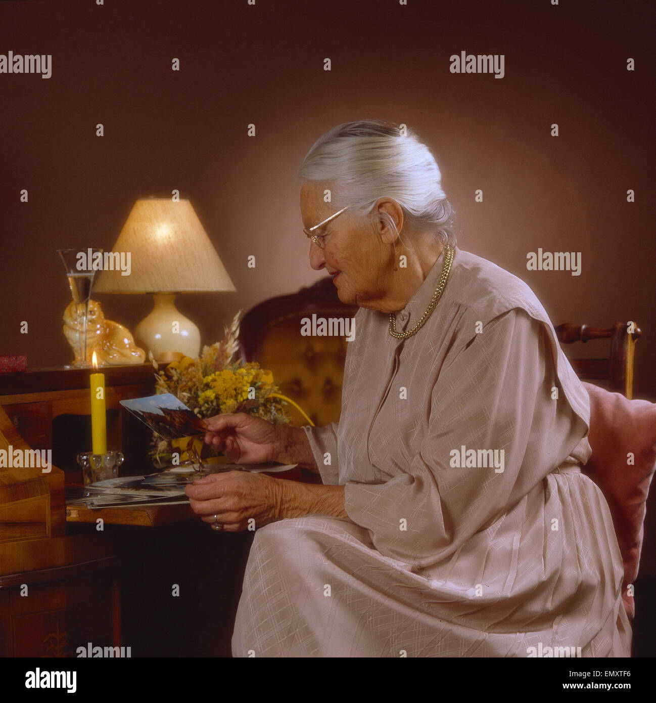 Seniorin mit Brille betrachtet Postkarte im Wohnzimmer, abends Stock Photo