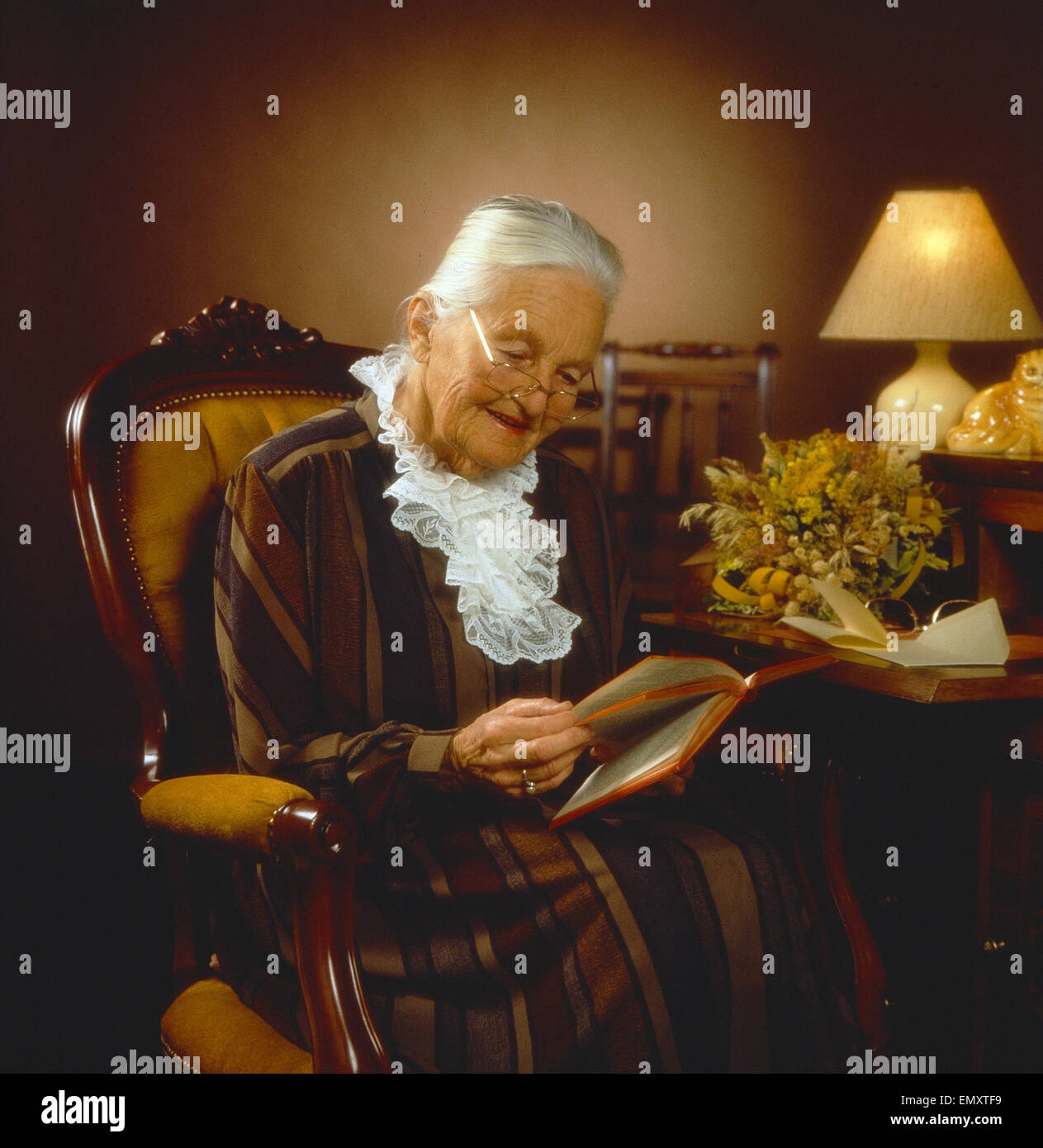 Seniorin sitzt in Sessel im Wohnzimmer und liest ein Buch, abends Stock Photo